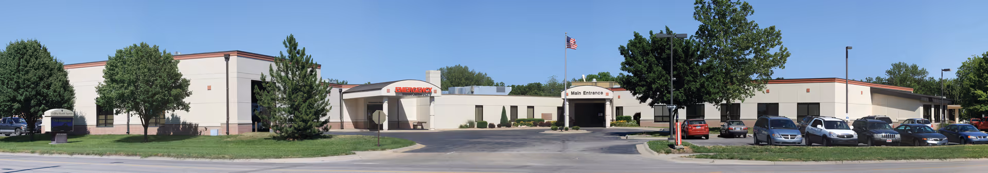 Panoramic exterior view of Coffey County Hospital building with a flagpole and parked cars.