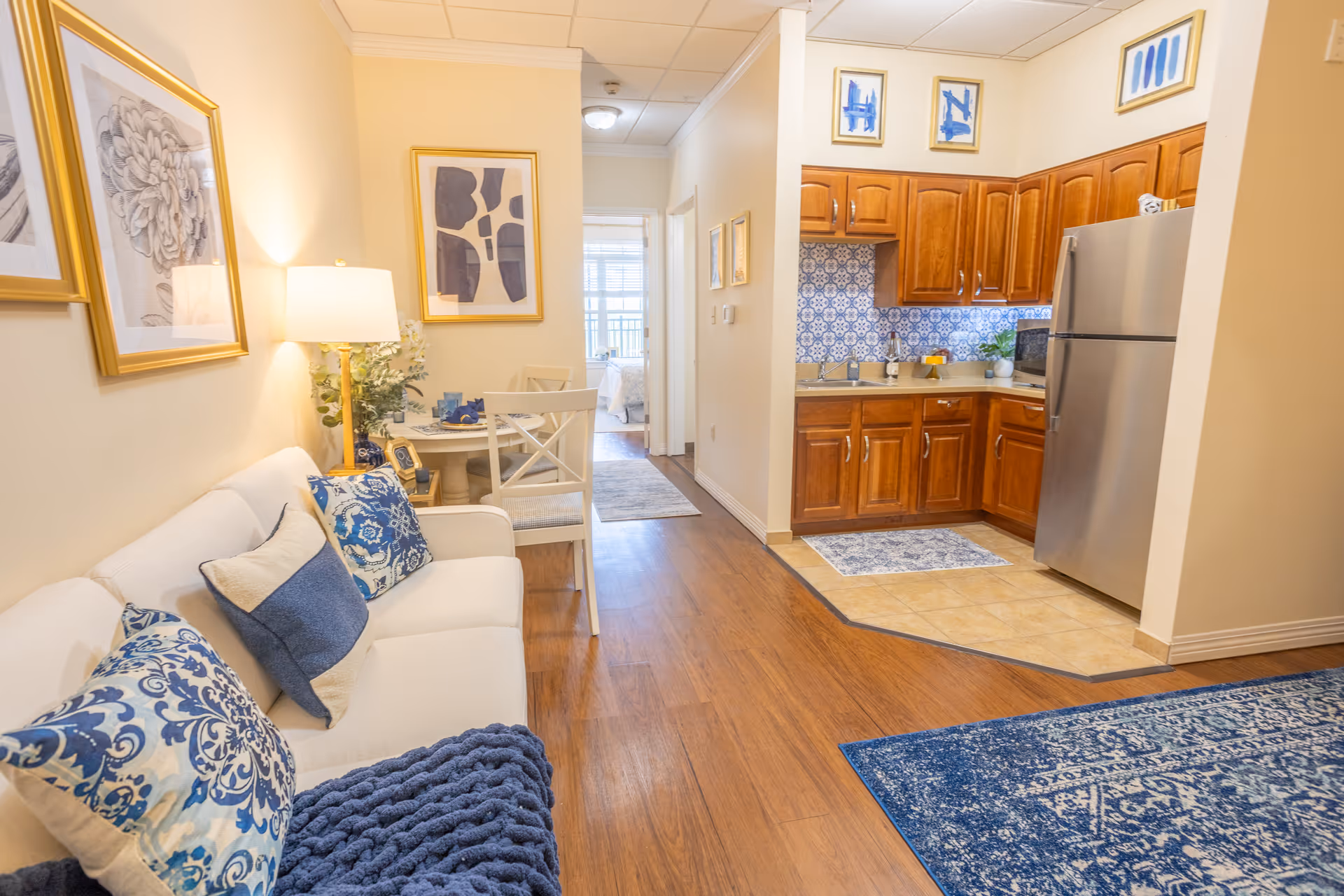 Bright living area with white sofa, blue cushions, dining table, and adjacent kitchen.