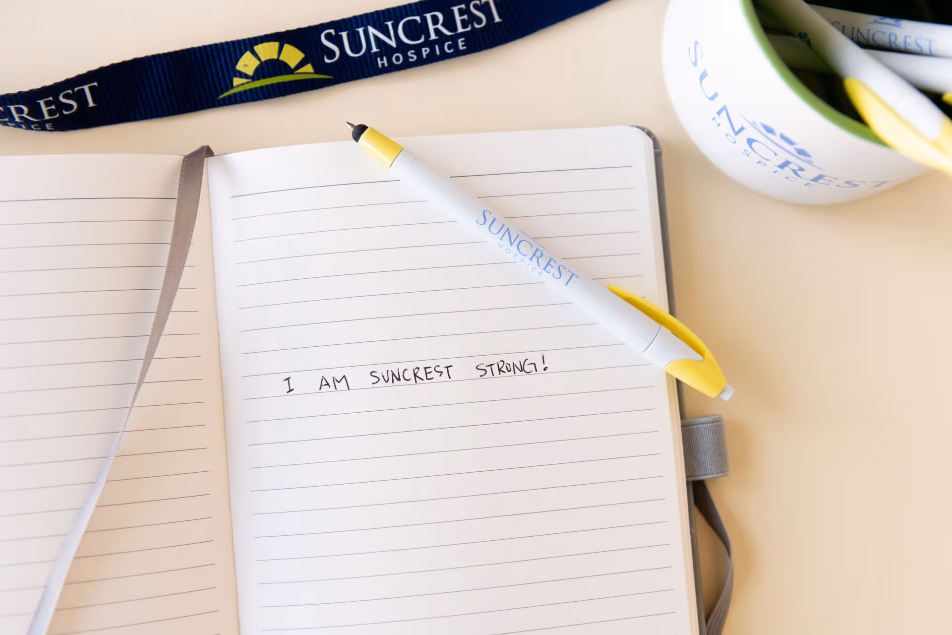 Notebook open on a desk with Suncrest Hospice lanyard, pen, and mug.