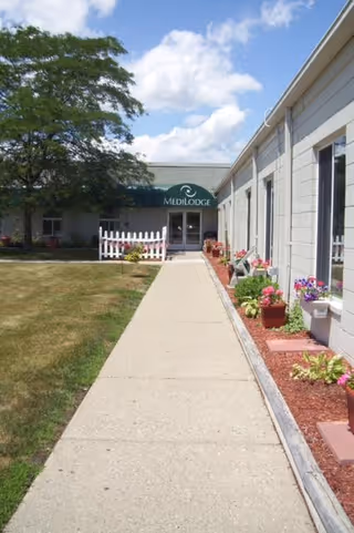 Sidewalk leading to the entrance of Medilodge of Richmond, with a green awning displaying the facility's name. The building is light-colored with windows and flower beds along the right side. A tree and grassy area are on the left under a partly cloudy sky.