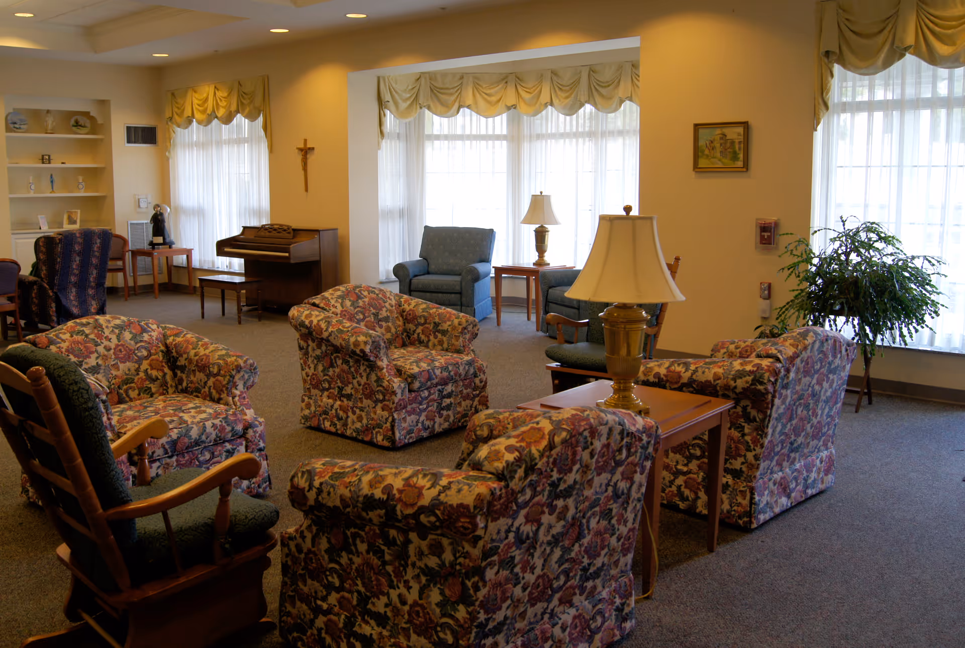 A cozy living room area in a memory care center with floral upholstered armchairs arranged around wooden side tables with lamps. The room has large windows with sheer curtains and valances, a piano against the wall, a rocking chair, and a potted plant near the window. The walls are decorated with a crucifix and a framed picture.