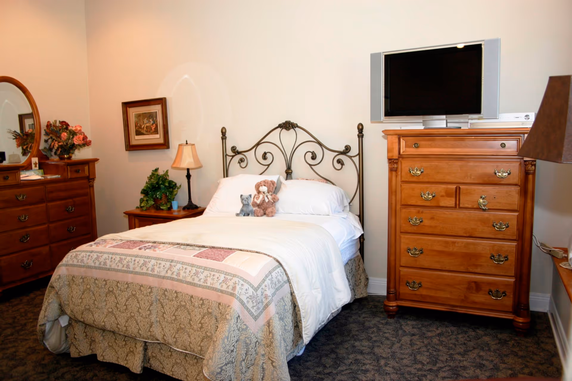 Cozy bedroom with a neatly made bed topped with stuffed animals, an ornate metal headboard, wooden dressers, a TV, and lamps.