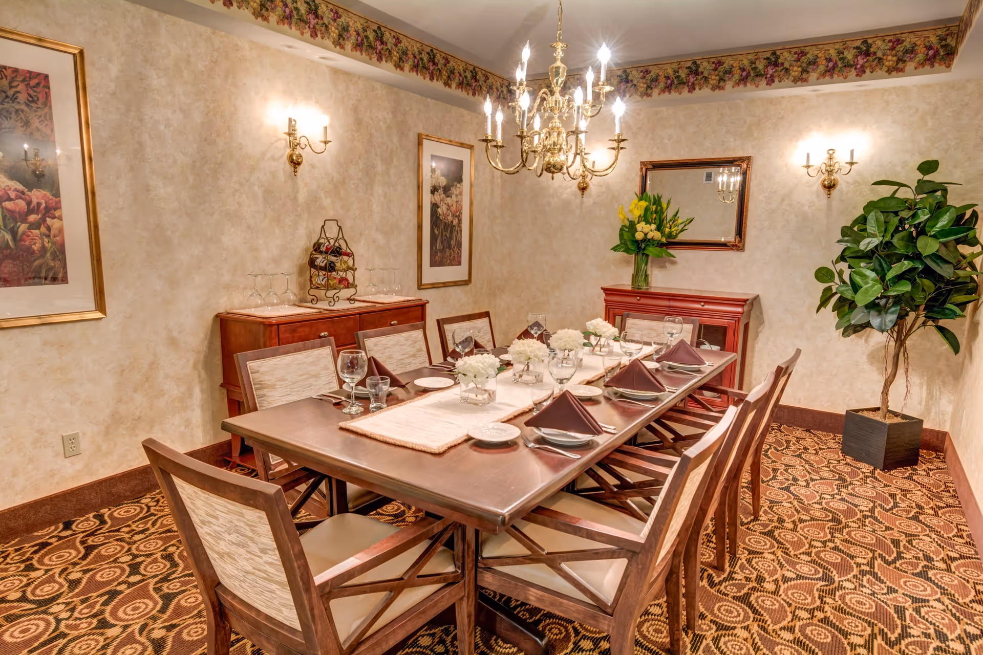 A formal dining room with a long wooden table set for eight people. The table is decorated with white floral centerpieces and brown folded napkins. The room features patterned carpet, beige textured walls with floral border trim near the ceiling, two framed floral paintings, a large mirror above a wooden sideboard with a vase of yellow flowers, and a potted plant in the corner. A brass chandelier and wall sconces provide lighting.