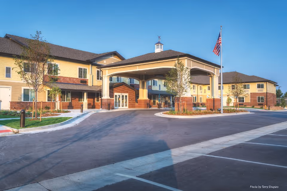 A modern assisted living facility exterior with a covered entrance and American flag.