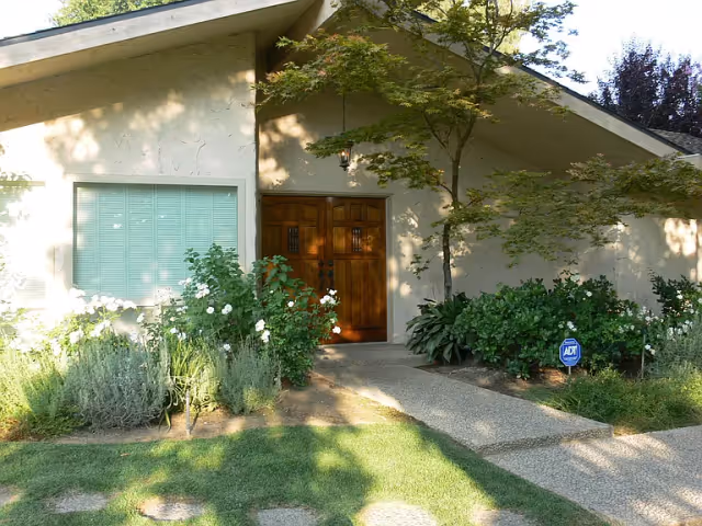 Front entrance of a single-story house with wooden double doors, a window with blinds, and a landscaped front yard with shrubs and a walkway.