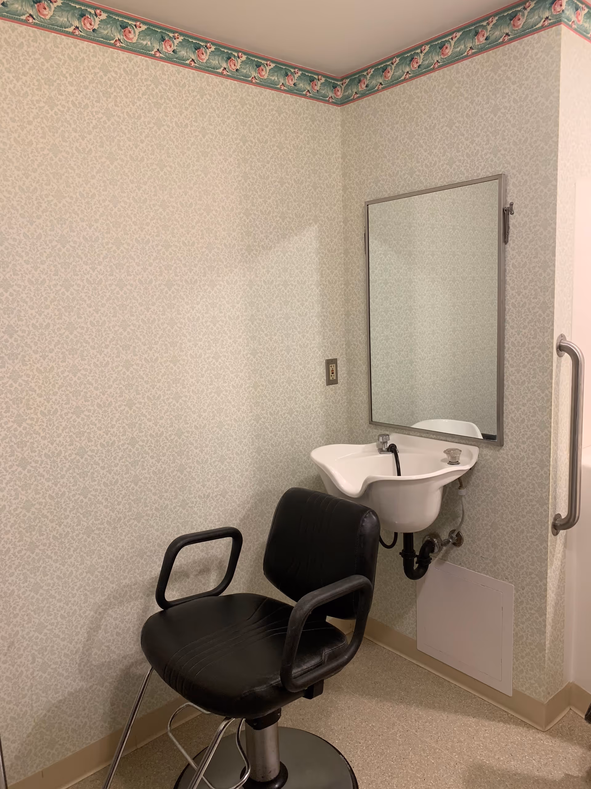 A small room with a black salon chair in front of a white wall-mounted sink designed for hair washing. There is a rectangular mirror above the sink and a floral wallpaper border near the ceiling. A metal grab bar is visible on the right side of the image.