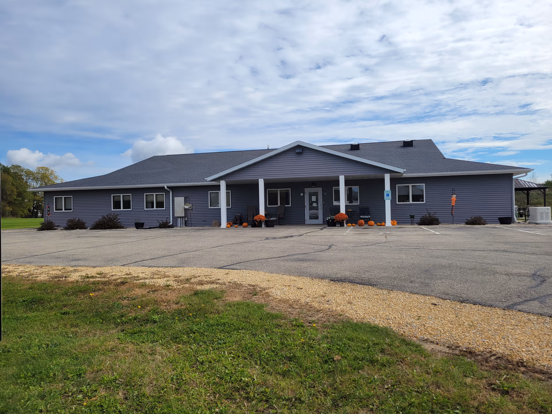 Single-story building with gray siding and a dark roof, featuring a covered entrance with white pillars. The front area has several pumpkins and potted plants, with a paved parking lot and green grass in the foreground under a partly cloudy sky.