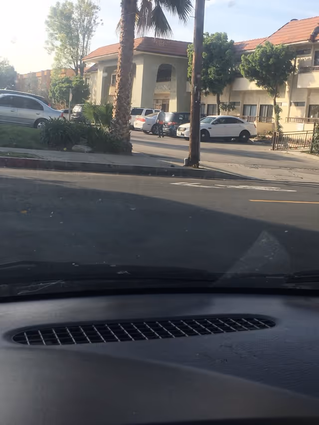 View from inside a car looking out through the windshield at the front exterior of a two-story building with a red-tiled roof, palm trees, parked cars, and a person standing near the entrance.
