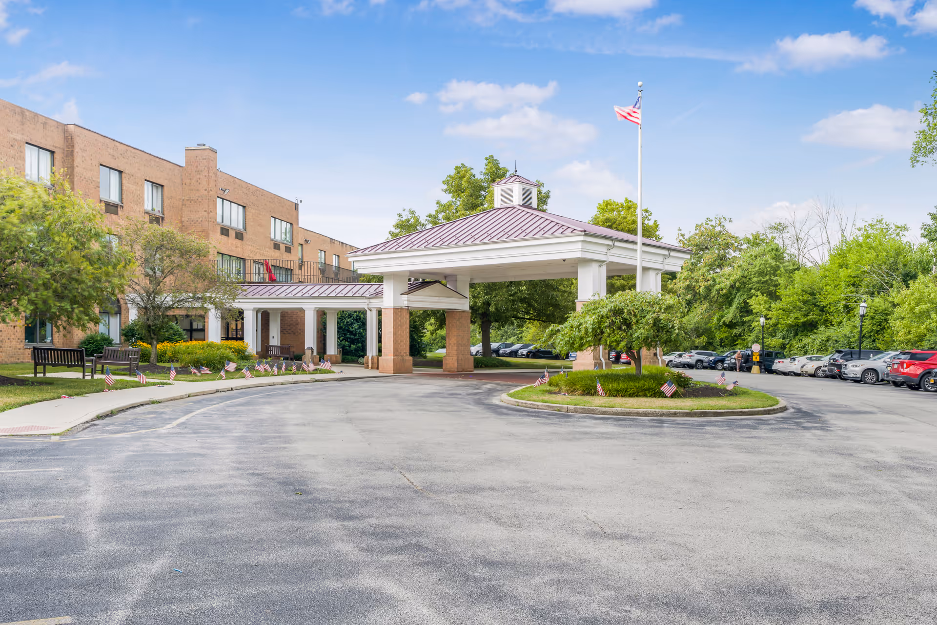 Front entrance with a covered porte-cochère at a brick rehabilitation and healthcare center, an American flagpole, and a surrounding parking lot.