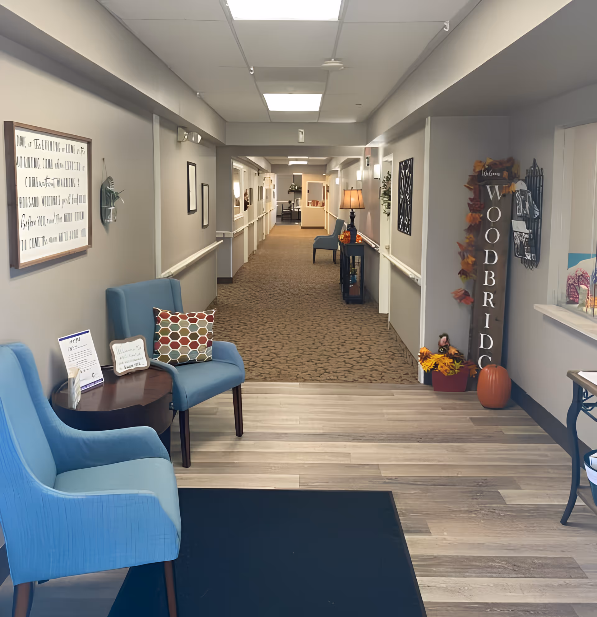 Well-lit assisted living hallway and seating area with blue chairs, decorative pillows, a lamp, and a vertical "Woodbridge" sign with fall decorations.