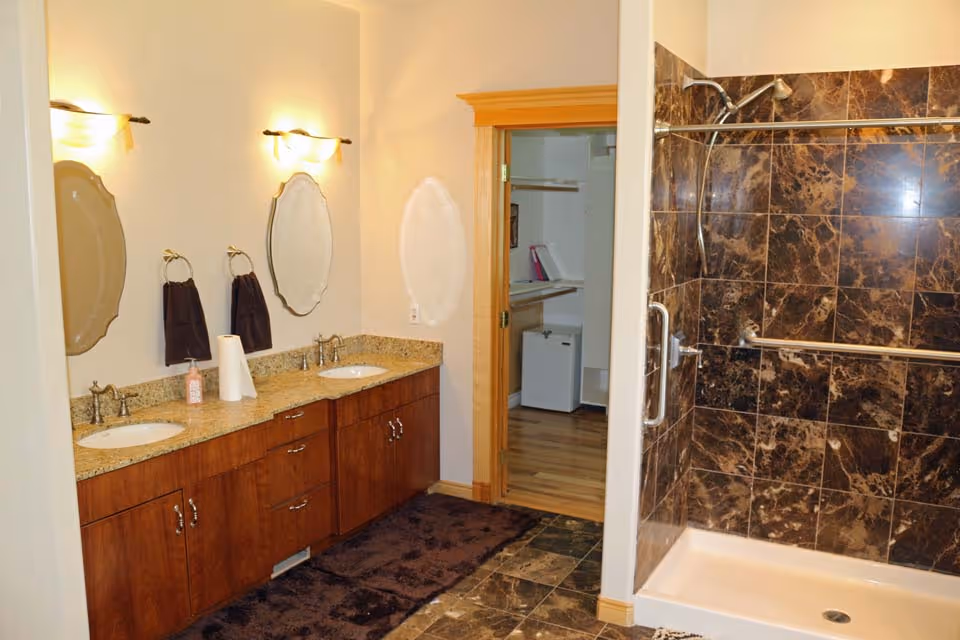 Spacious bathroom with a double-sink wooden vanity and granite countertop beside a dark-tiled walk-in shower.