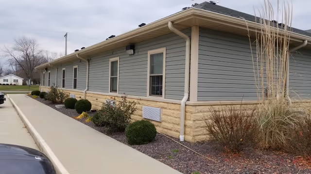 Exterior side view of a single-story building with light blue siding and beige stone foundation, featuring several windows and a sidewalk with landscaped bushes and plants alongside it.