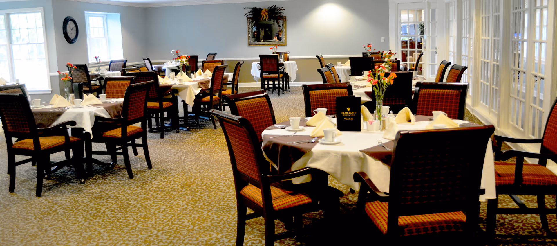 A well-lit dining room with multiple tables covered in white and brown tablecloths, each set with napkins, cups, and small flower arrangements. The room has patterned carpet flooring, large windows letting in natural light, and a clock on the wall. Chairs with red and black upholstery surround the tables.