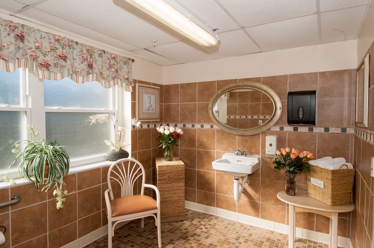 Well-lit tiled restroom with a wall-mounted sink and oval mirror, a chair, plants, and a basket of towels.