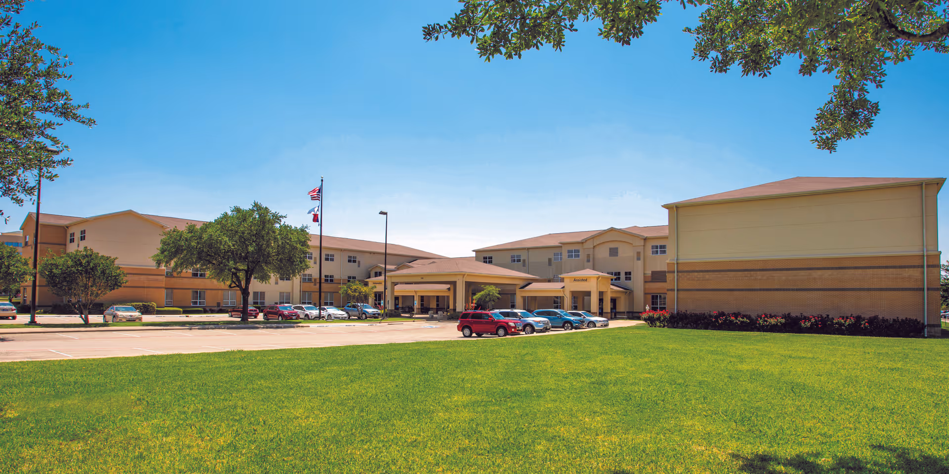 Front exterior of a multi-story senior living building with a wide green lawn, parked cars, and a clear blue sky.