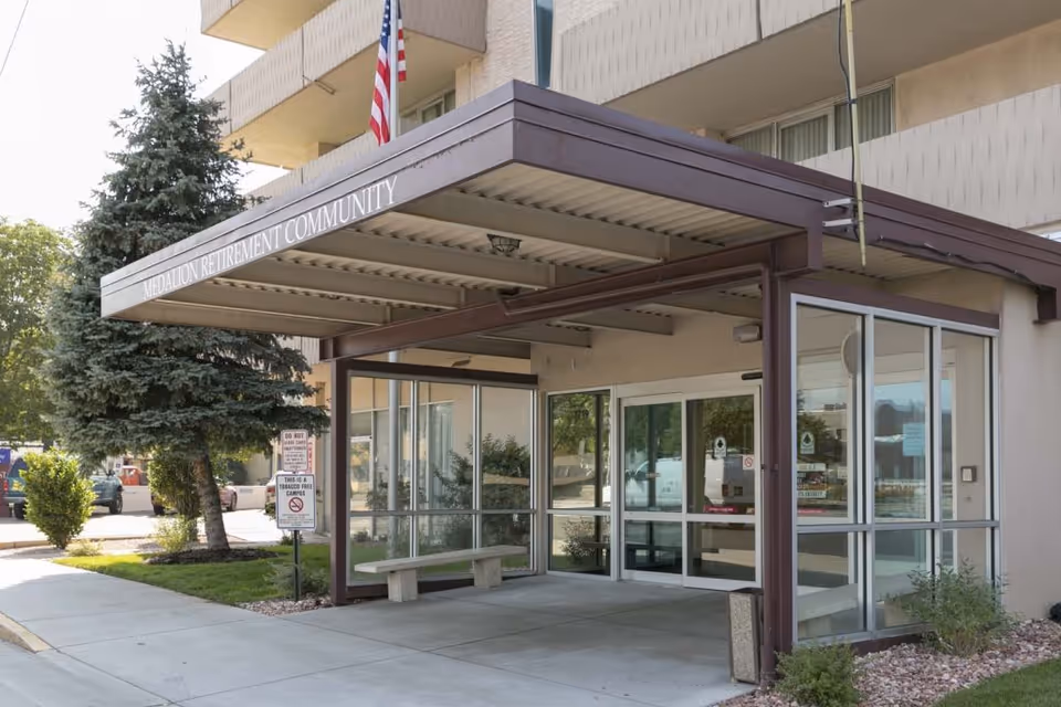 Covered entrance with glass doors and a bench at the Medallion Retirement Community building, with an American flag overhead.