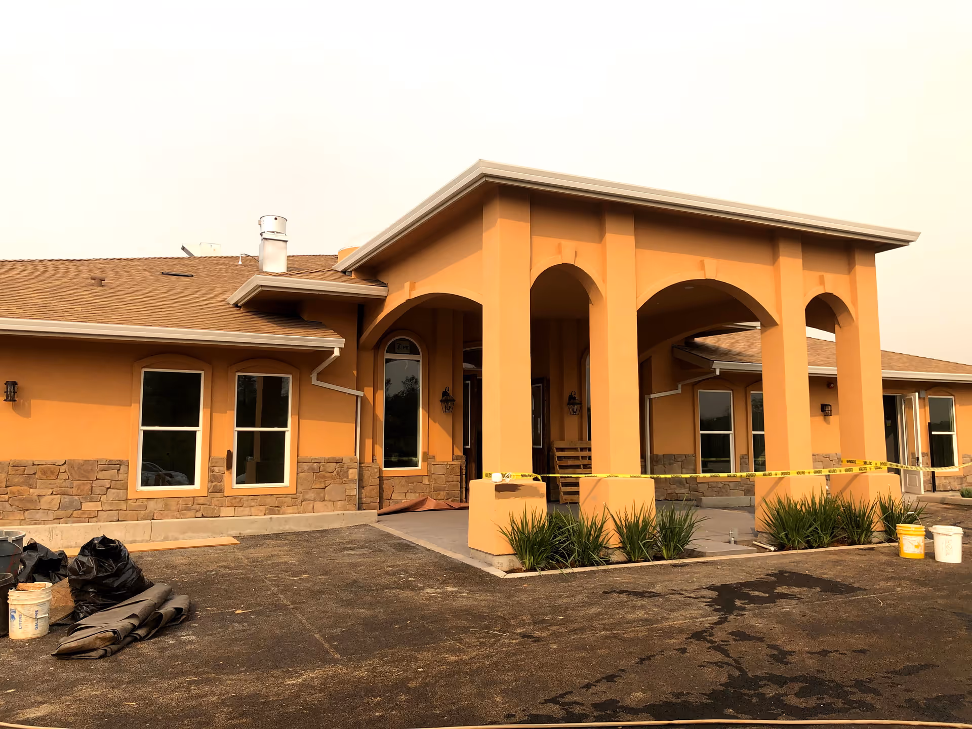 Front entrance of a peach-colored senior living building with arched portico, landscaping, and construction materials with caution tape.