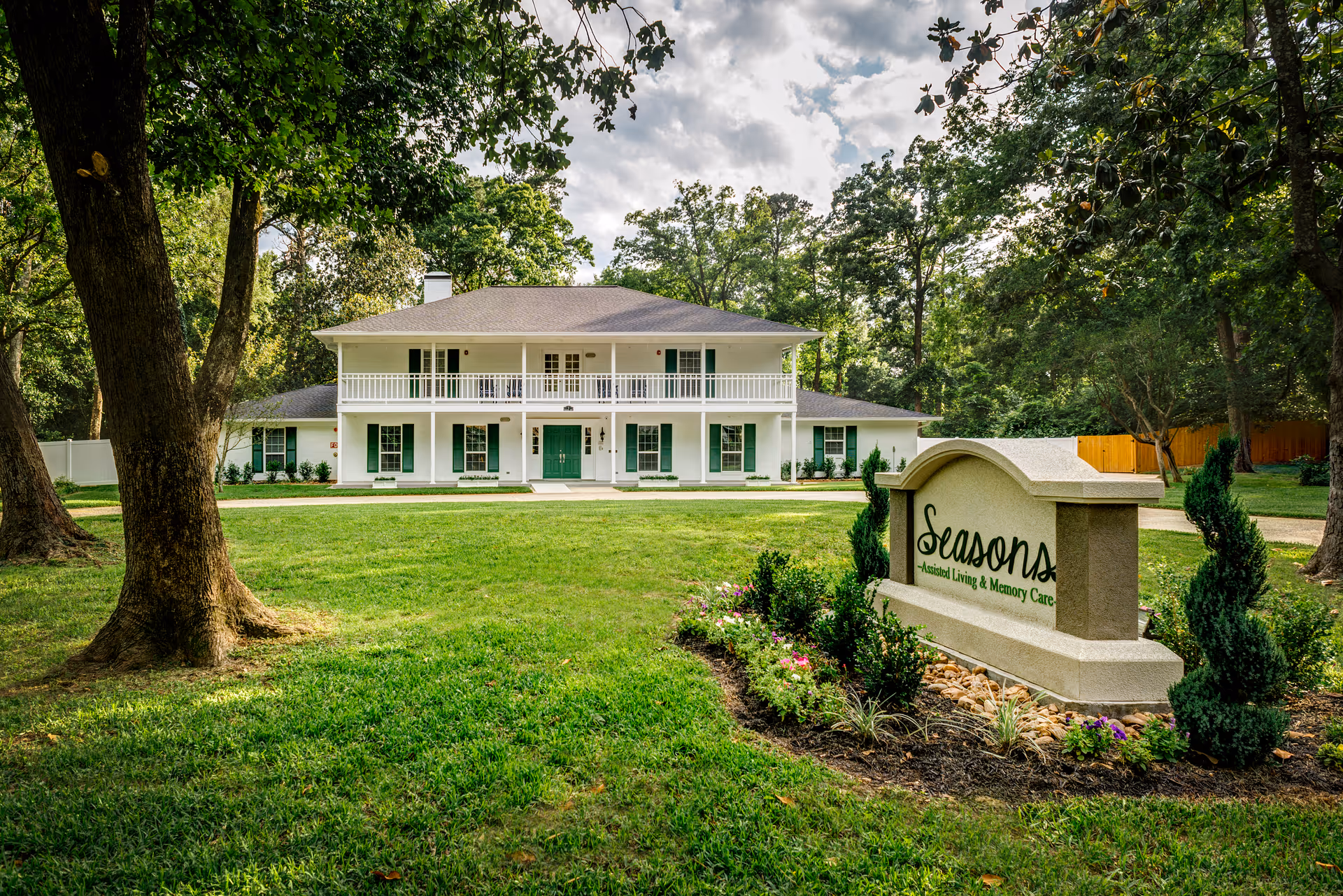 A large two-story white building with green shutters and a green front door, surrounded by trees and a well-maintained lawn. In the foreground, there is a landscaped sign that reads 'Seasons Assisted Living & Memory Care'.