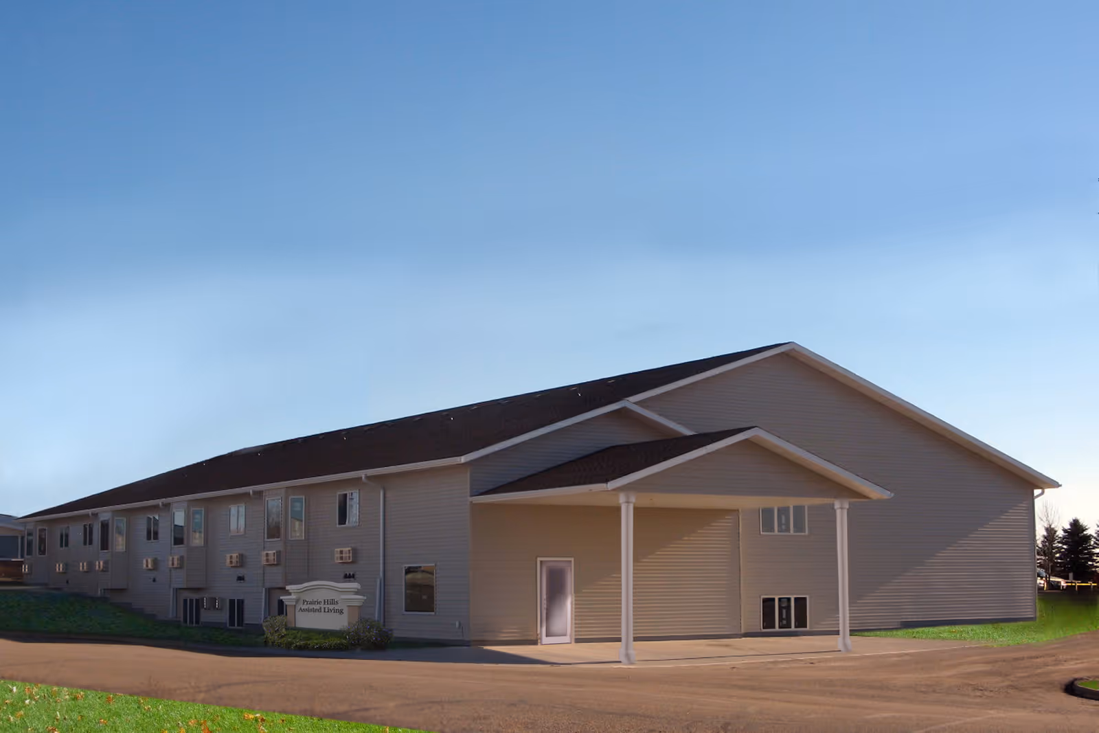 Exterior view of a single-story assisted living facility building with beige siding, a dark roof, and a covered entrance supported by white columns. The building is surrounded by a paved driveway and some grass, under a clear blue sky.