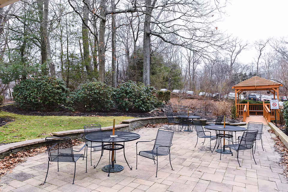 Outdoor patio area with several black metal tables and chairs on a paved surface. In the background, there is a wooden gazebo and leafless trees and bushes surrounding the area.