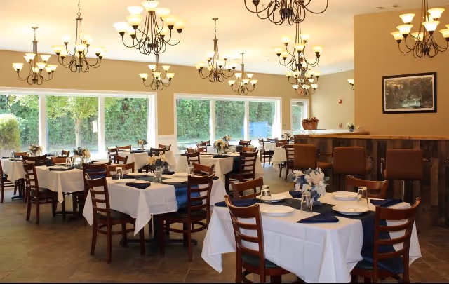 Dining room with white tablecloths, wooden chairs, chandeliers, and large windows in a senior living facility.