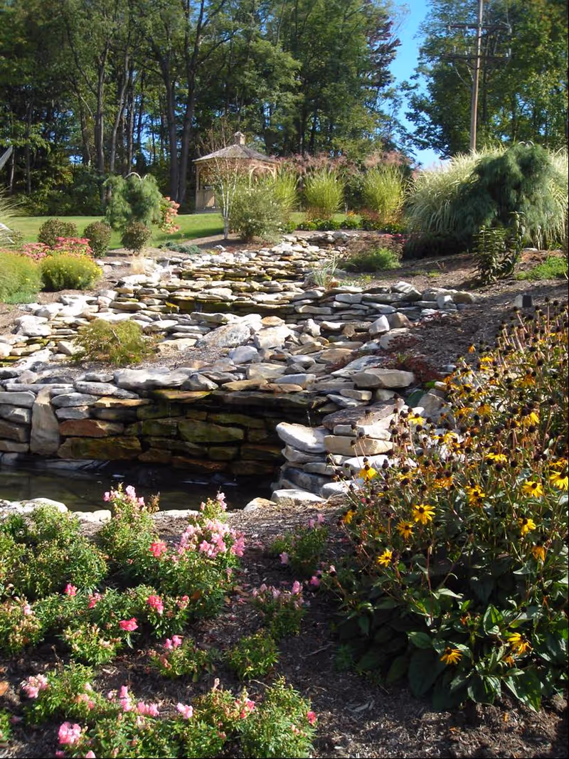 Well-manicured garden with a stacked rock waterfall, pond, and gazebo in a sunny outdoor setting.