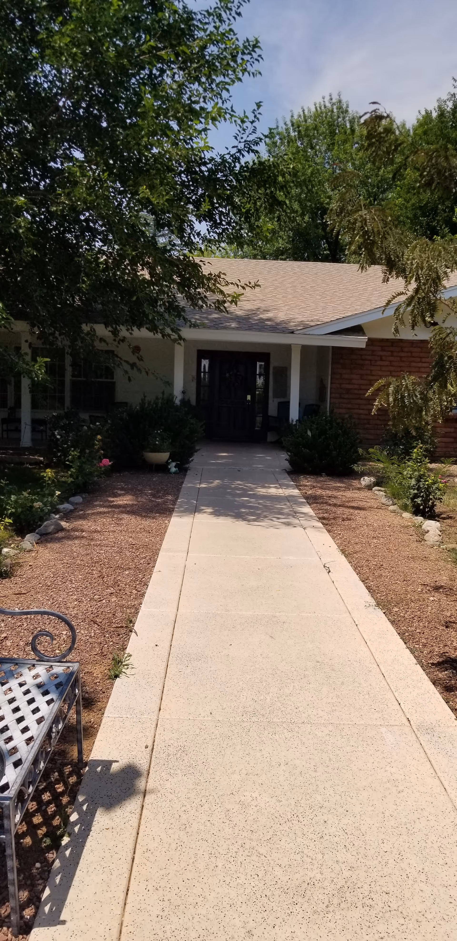 A concrete walkway leading to the front entrance of a single-story building with a covered porch. The walkway is bordered by landscaped areas with small bushes, plants, and trees. A metal bench is visible on the left side of the walkway. The sky is clear with some clouds.