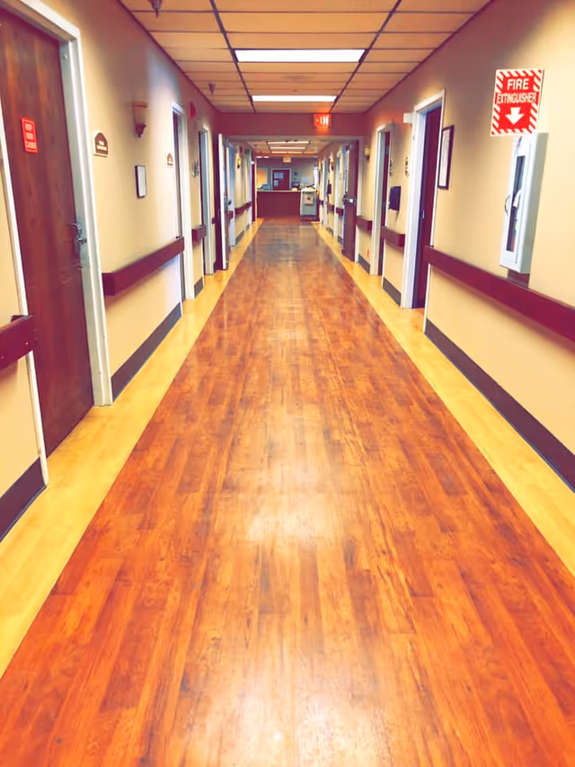 A long, clean hallway in a rehabilitation and healing center with wooden floors, beige walls, and multiple doors on both sides. Handrails run along the walls, and there is a fire extinguisher mounted on the right wall. The ceiling has fluorescent lights, and at the end of the hallway, there is a reception or nurse's station.