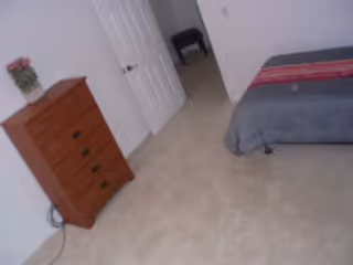 A bedroom with a bed, dresser, plant, and a doorway in a neutral-toned room.