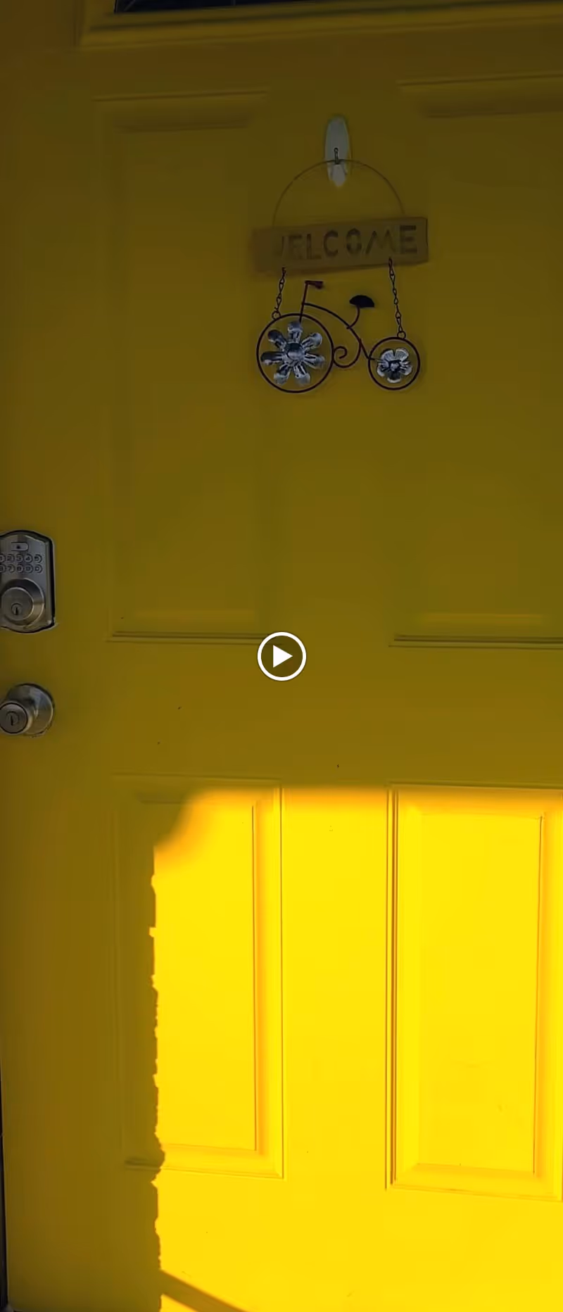 A bright yellow door with a silver keypad lock and handle on the left side. Hanging on the door is a decorative sign shaped like a bicycle with flower-shaped wheels and a wooden plaque above it that reads 'WELCOME'.