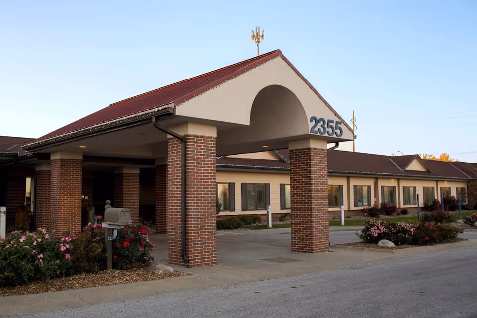 Exterior view of a single-story brick building with a covered entrance supported by brick columns. The building has a red roof and beige walls with multiple windows. The number 2355 is displayed prominently on the entrance structure. There are flower bushes and landscaping around the entrance area.