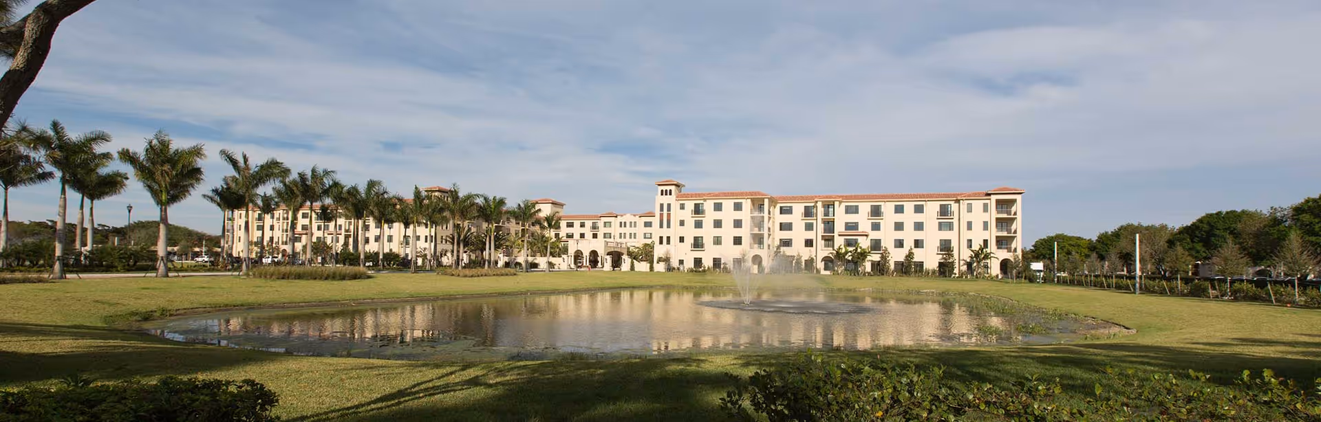 Exterior view of Candy Cohn senior living facility with a pond, lawn, and palm trees.