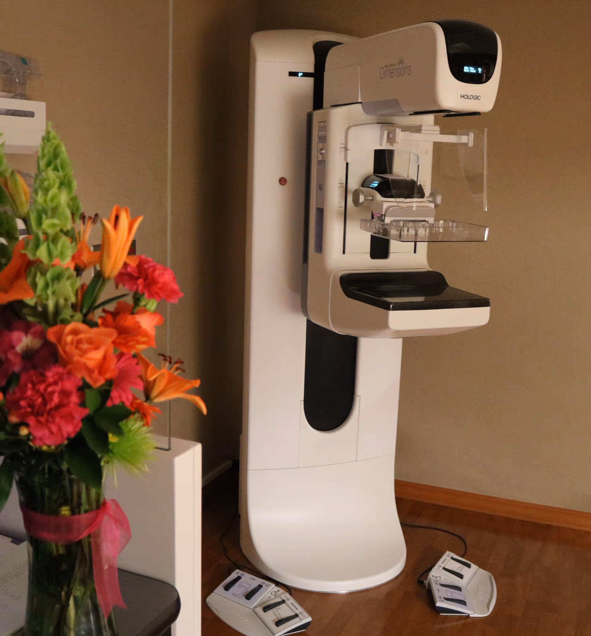 A white medical imaging machine stands in a clinical room beside a bouquet of flowers.