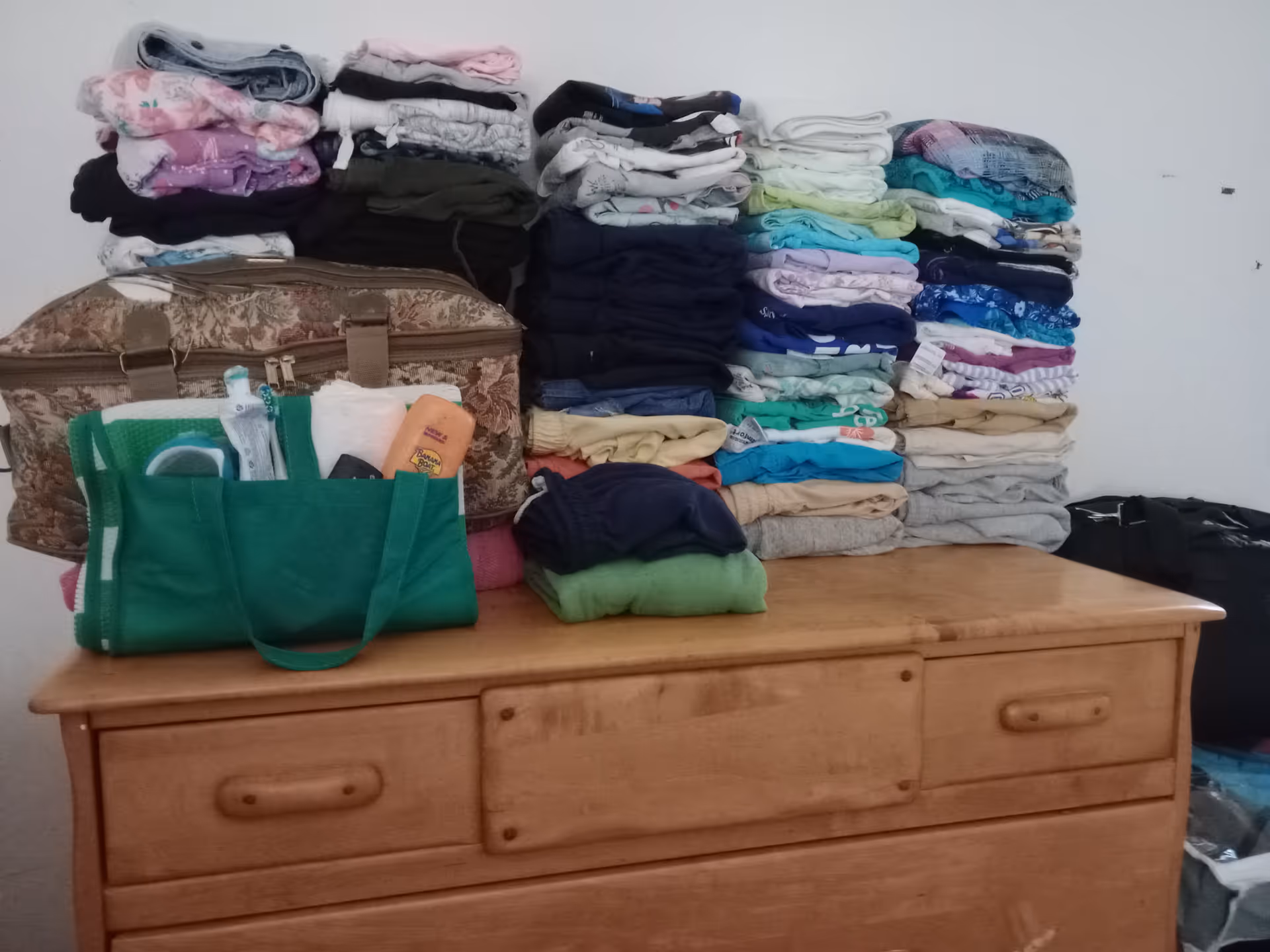 Stacks of folded clothes and a green tote on a wooden dresser.