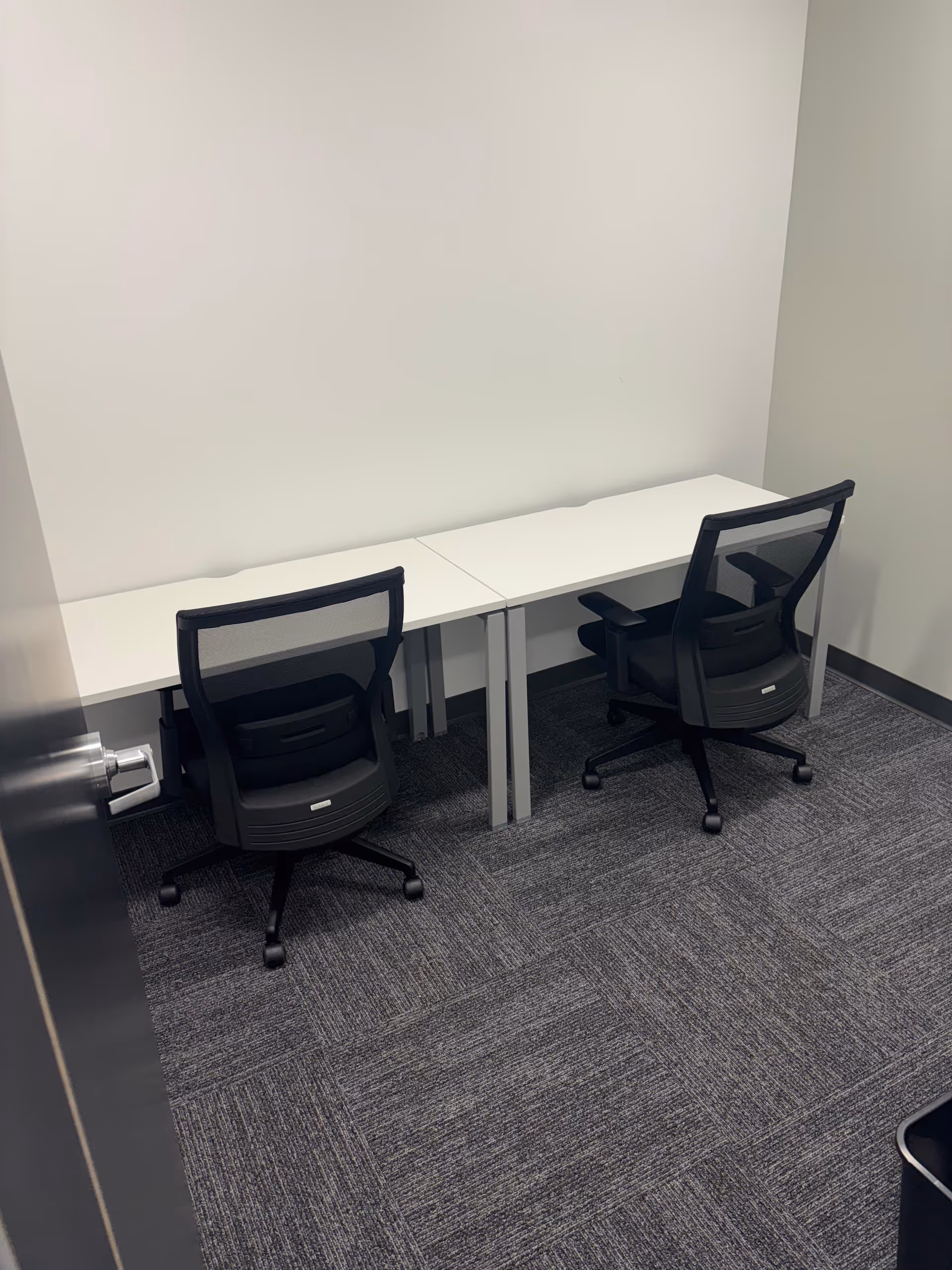 Two white desks with black office chairs against a blank white wall.