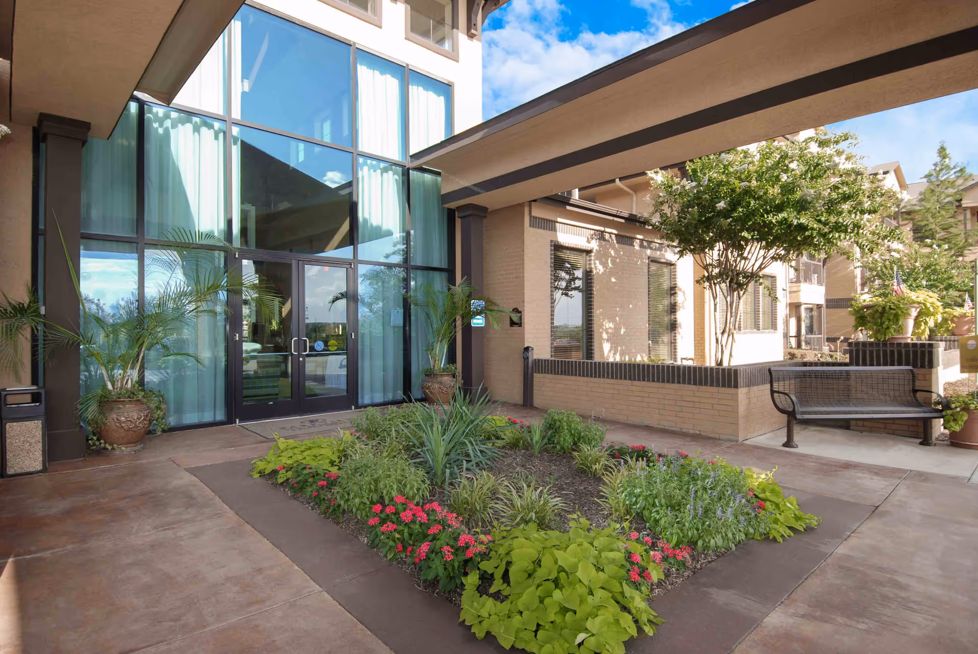 Entrance of a senior living facility with large glass doors and windows, surrounded by potted plants and a landscaped garden bed with various green plants and red flowers. There is a bench to the right and a covered walkway overhead. The building exterior is light-colored brick with black trim.