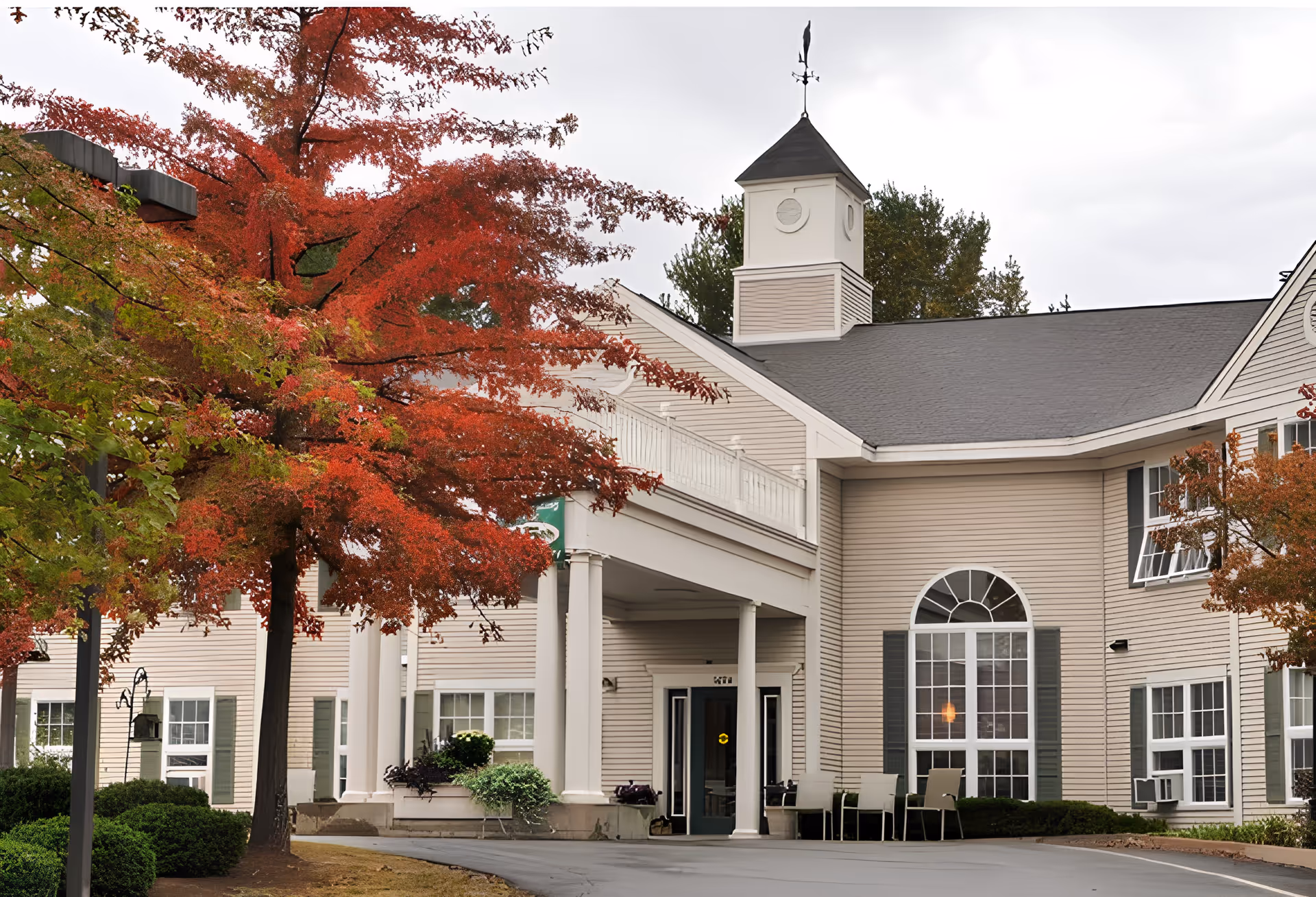 Exterior view of a senior living community building with beige siding, white columns, and a small tower with a weather vane on top. There are red and green trees in the foreground, and a paved driveway leading to the entrance with chairs placed outside.