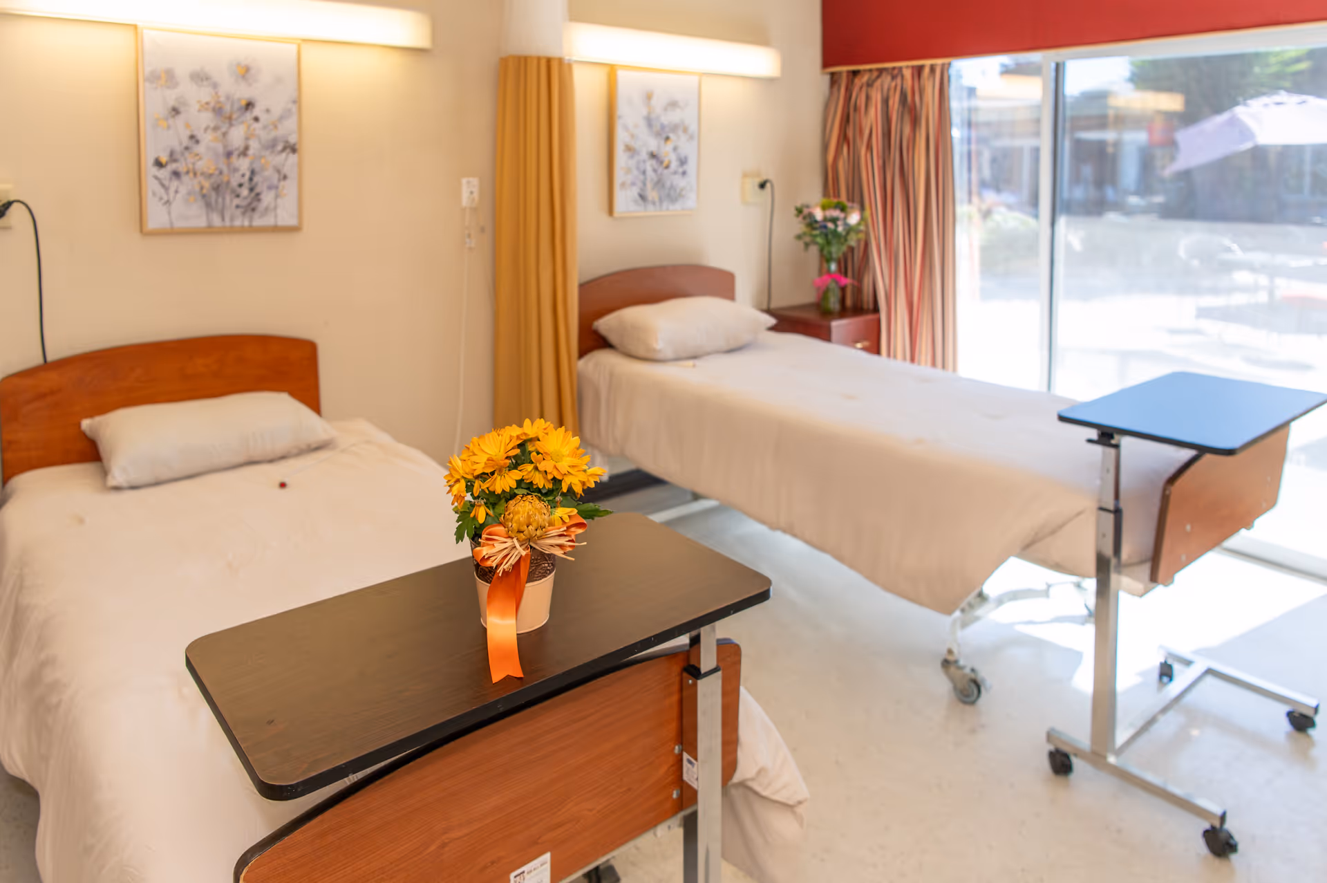 Bright patient room with two beds, a small table, and a vase of flowers.