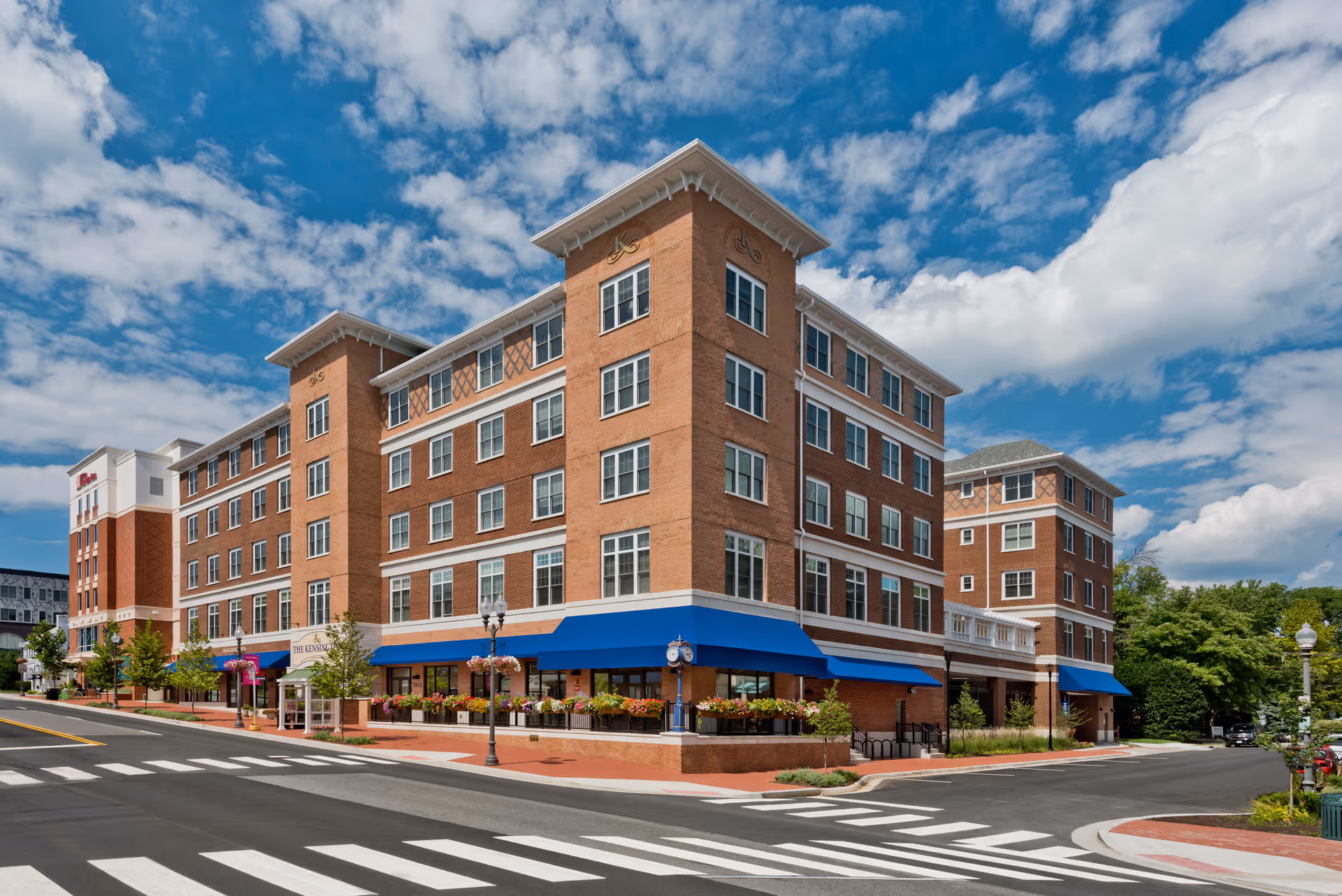 Five-story brick senior living building with blue awnings on a street corner under a partly cloudy sky.
