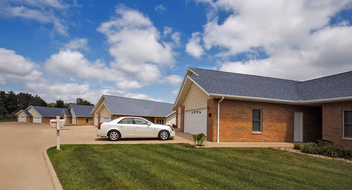 Row of brick senior living buildings with garages, a parked car, green lawn, under blue sky