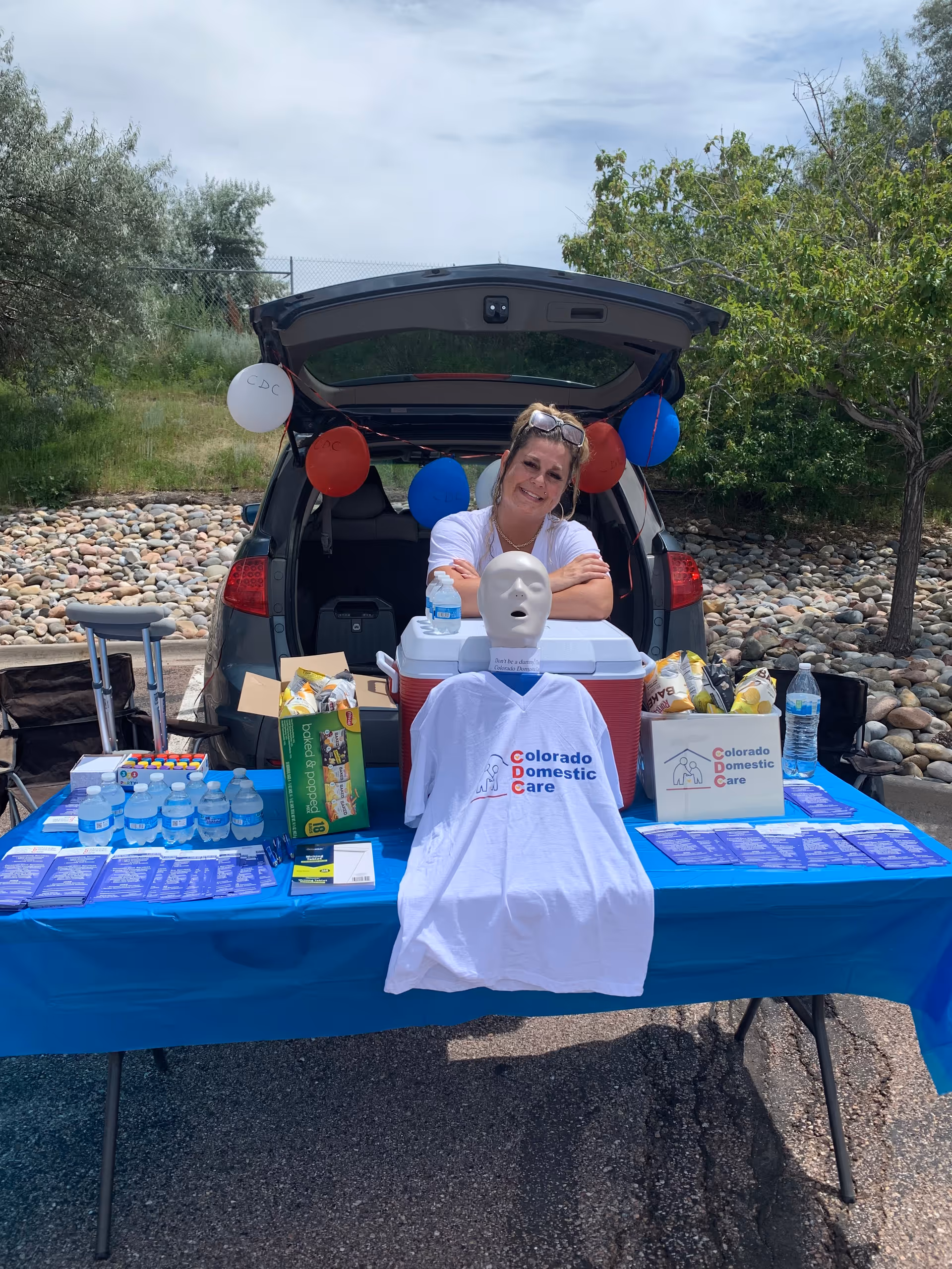 Staff member promoting Colorado Domestic Care at an outdoor table with balloons and promotional materials.