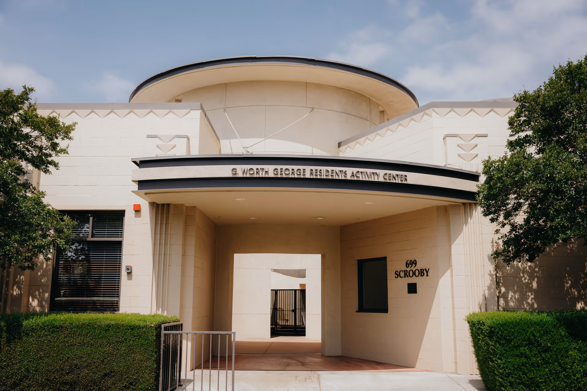 Front entrance of a beige residents' activity center with an arched canopy, signage reading '699 SCROOBY', and trimmed hedges.