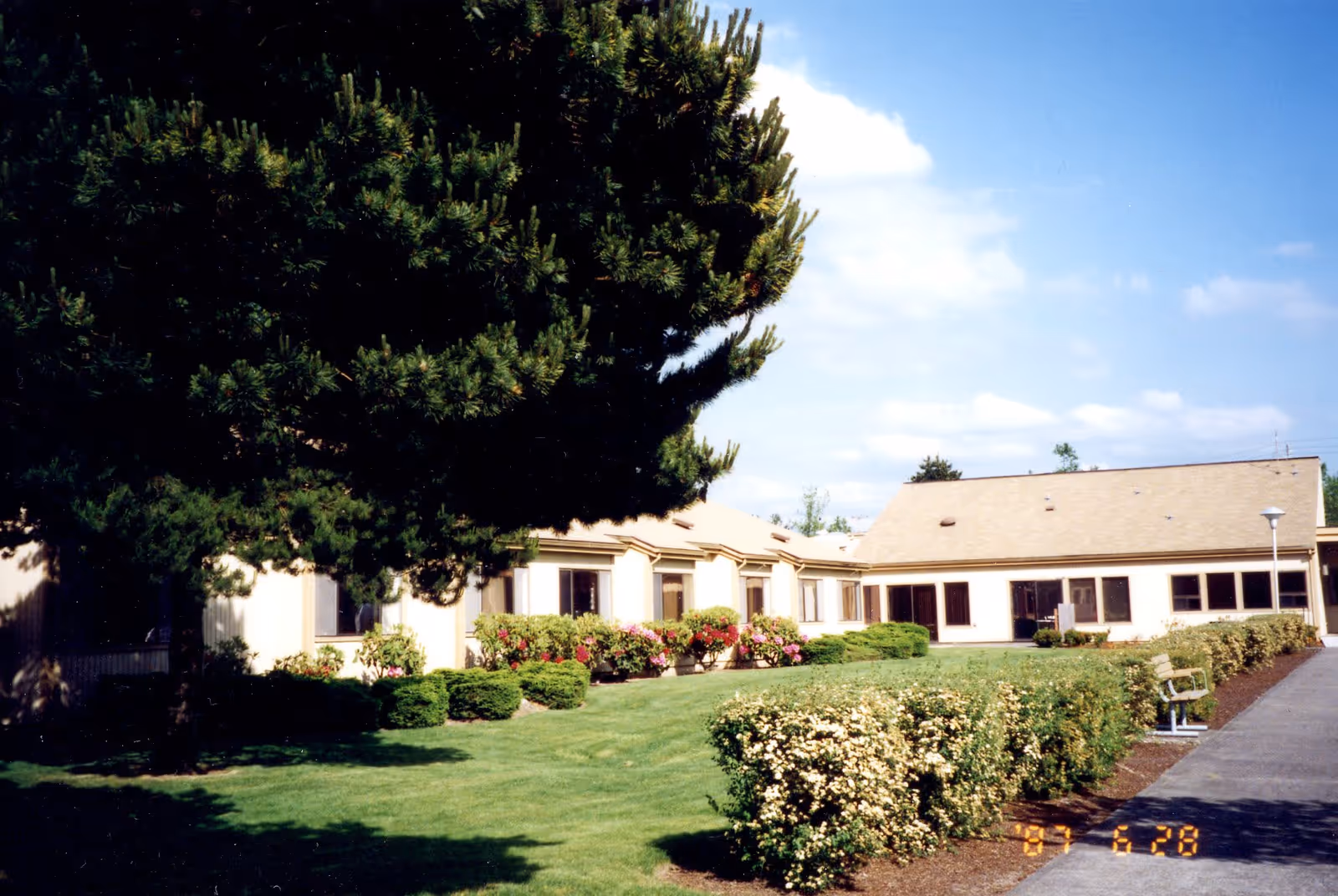 Single-story retirement building with a manicured lawn, shrubs, benches and a large tree under a blue sky.