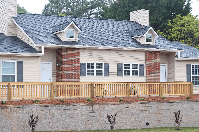 Exterior view of a single-story residential building with beige siding, dark gray roof shingles, and brick chimney accents. The building has multiple windows with dark shutters and two white doors. A wooden railing runs along a raised patio area in front of the building, with a stone retaining wall and small plants in the landscaped area below.