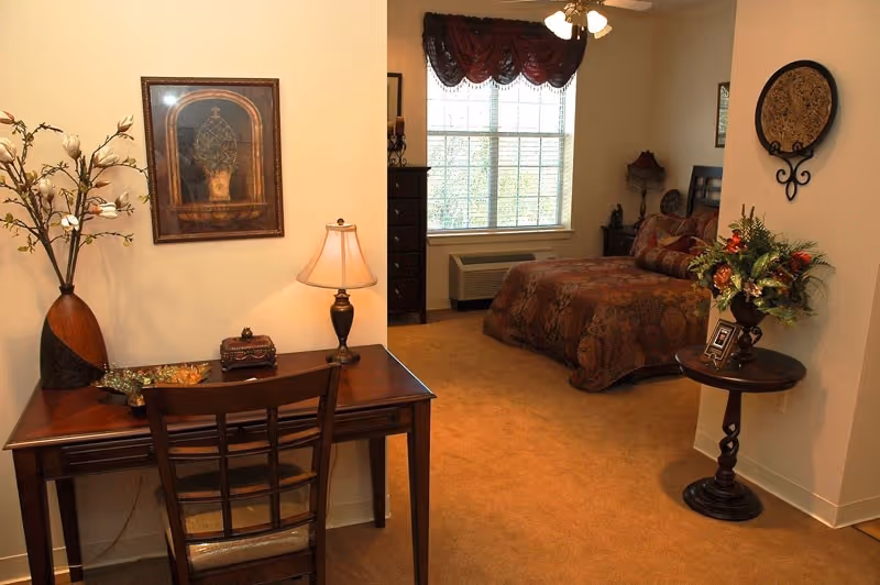 A cozy bedroom in an assisted living facility featuring a bed with patterned bedding, a window with burgundy valance curtains, a wooden dresser, and a small round table with a floral arrangement. In the foreground, there is a wooden desk with a chair, a table lamp, a decorative vase with flowers, and a framed picture on the wall.