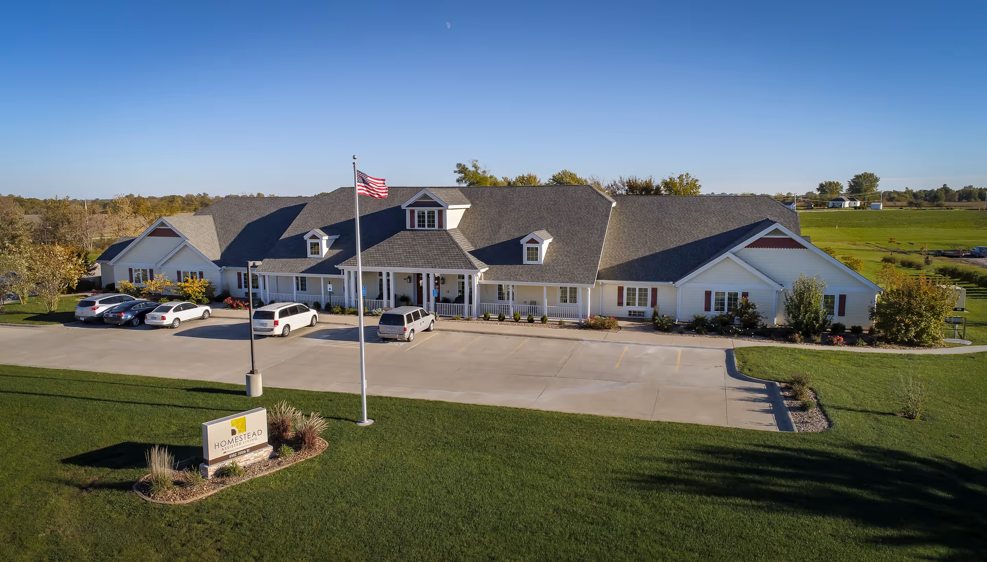 Front exterior of Homestead Assisted Living building with an American flag, parked cars, and a landscaped lawn.