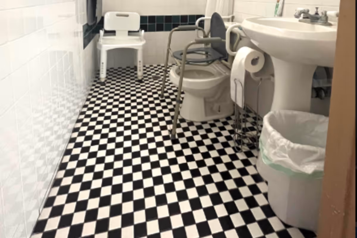 Accessible bathroom in senior facility with checkered tile floor, toilet, sink, and chair.