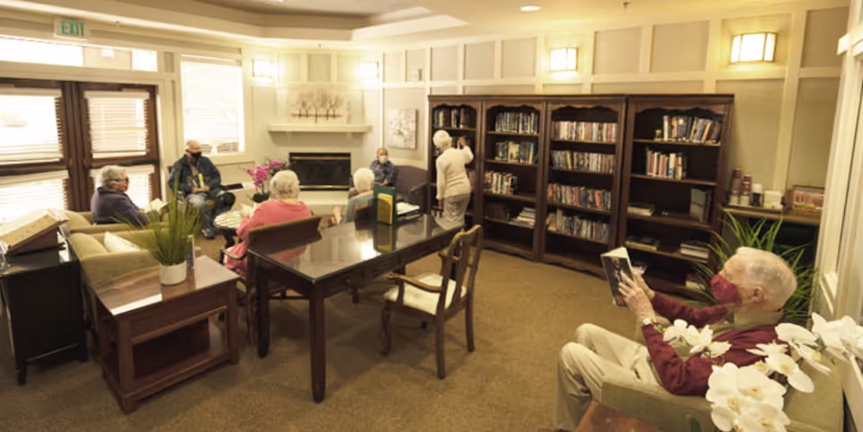 Several residents wearing masks sit and read in a comfortable communal library/lounge with bookshelves, a fireplace, and tables.