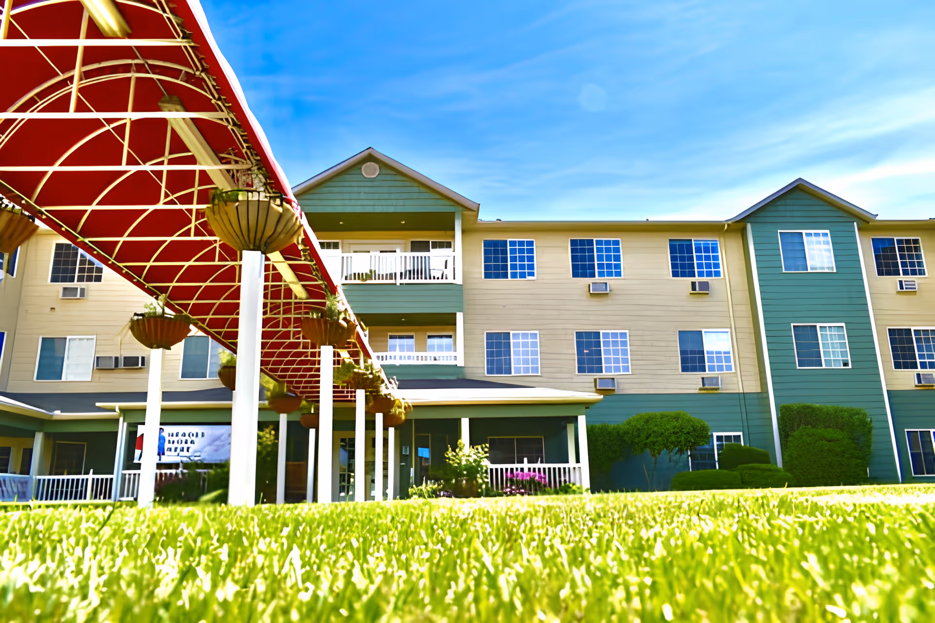 Exterior view of a multi-story senior living facility building with green and beige siding, multiple windows, and balconies. In the foreground, there is a well-maintained green lawn and a covered walkway with a red canopy and hanging flower pots. The sky is clear and blue.