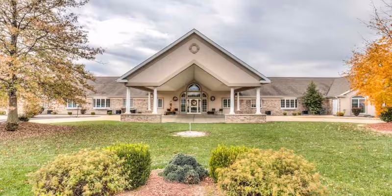 Front exterior view of Brookstone Estates of Effingham, a single-story building with a large covered entrance supported by white columns. The building is surrounded by green grass, bushes, and trees with autumn foliage under a cloudy sky.